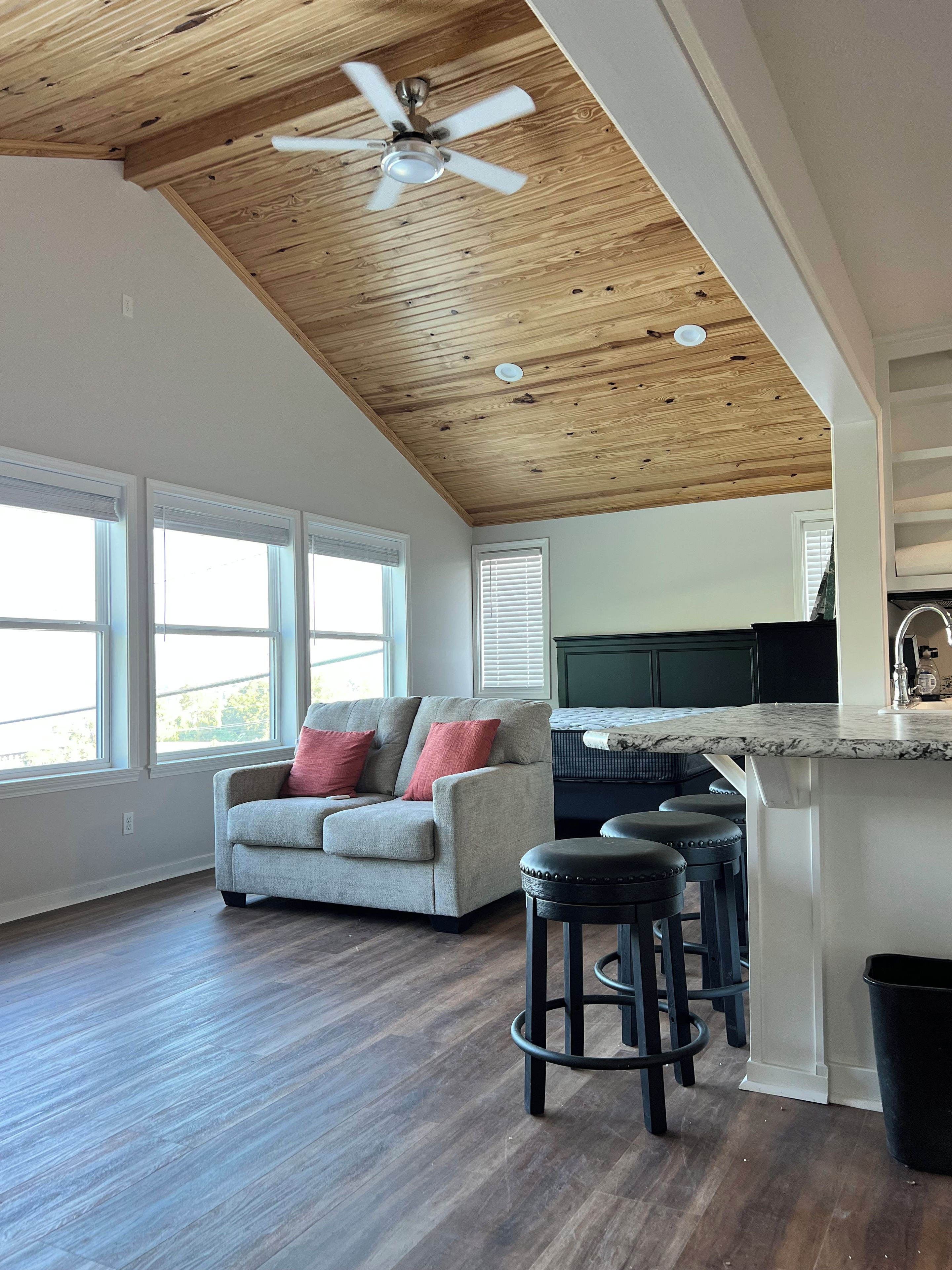 A spacious living area with a wooden ceiling, a grey sofa with red cushions, a kitchen island, and a bar stool.