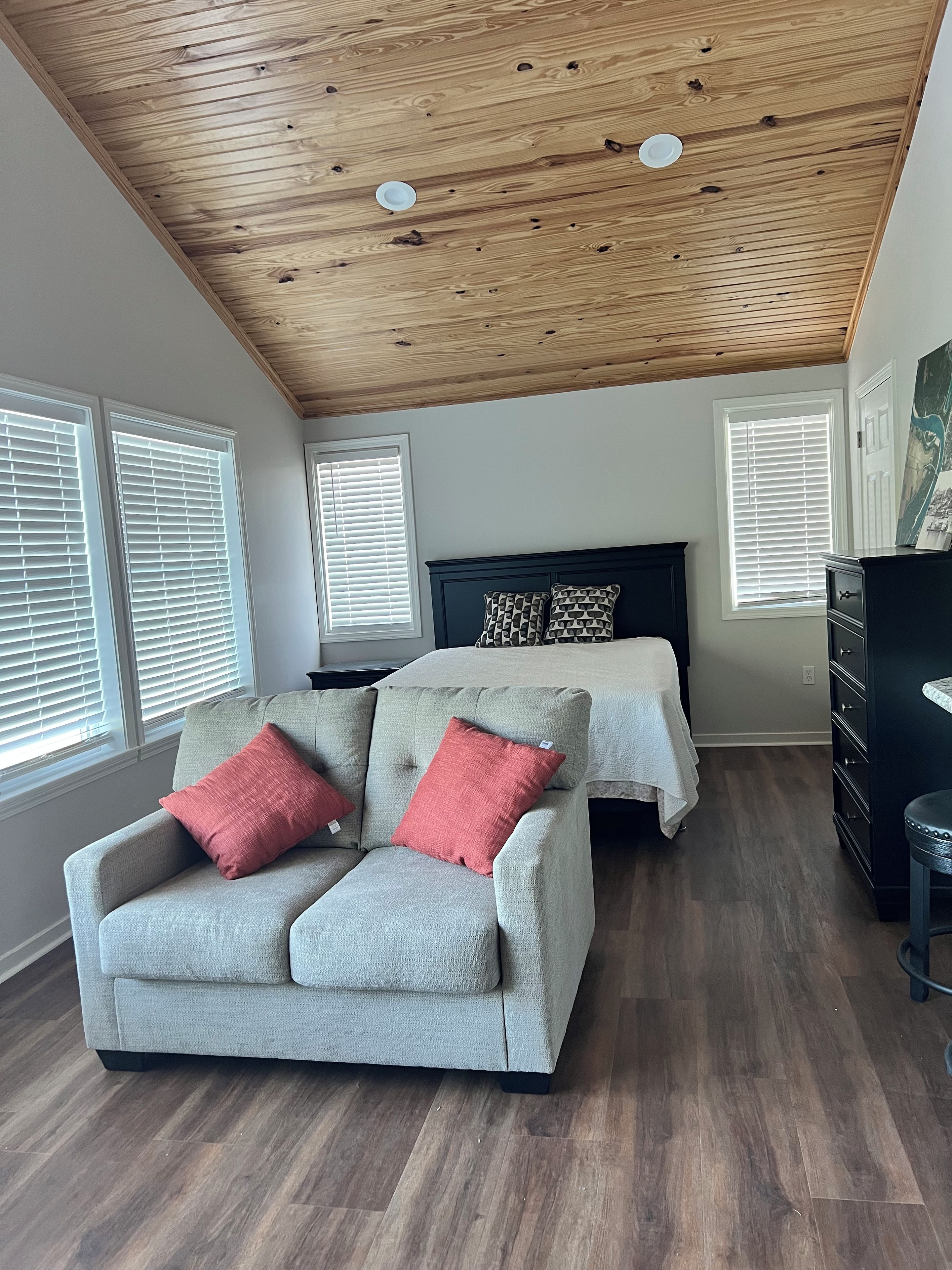 Una habitación con techo de madera, un sofá gris con cojines rojos, una cama con almohadones estampados y un aparador.