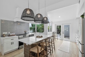 A modern kitchen with a large island, white cabinetry, and a black hanging light fixture. - Bridge-Way Villas at Pelican Bay (Naples)