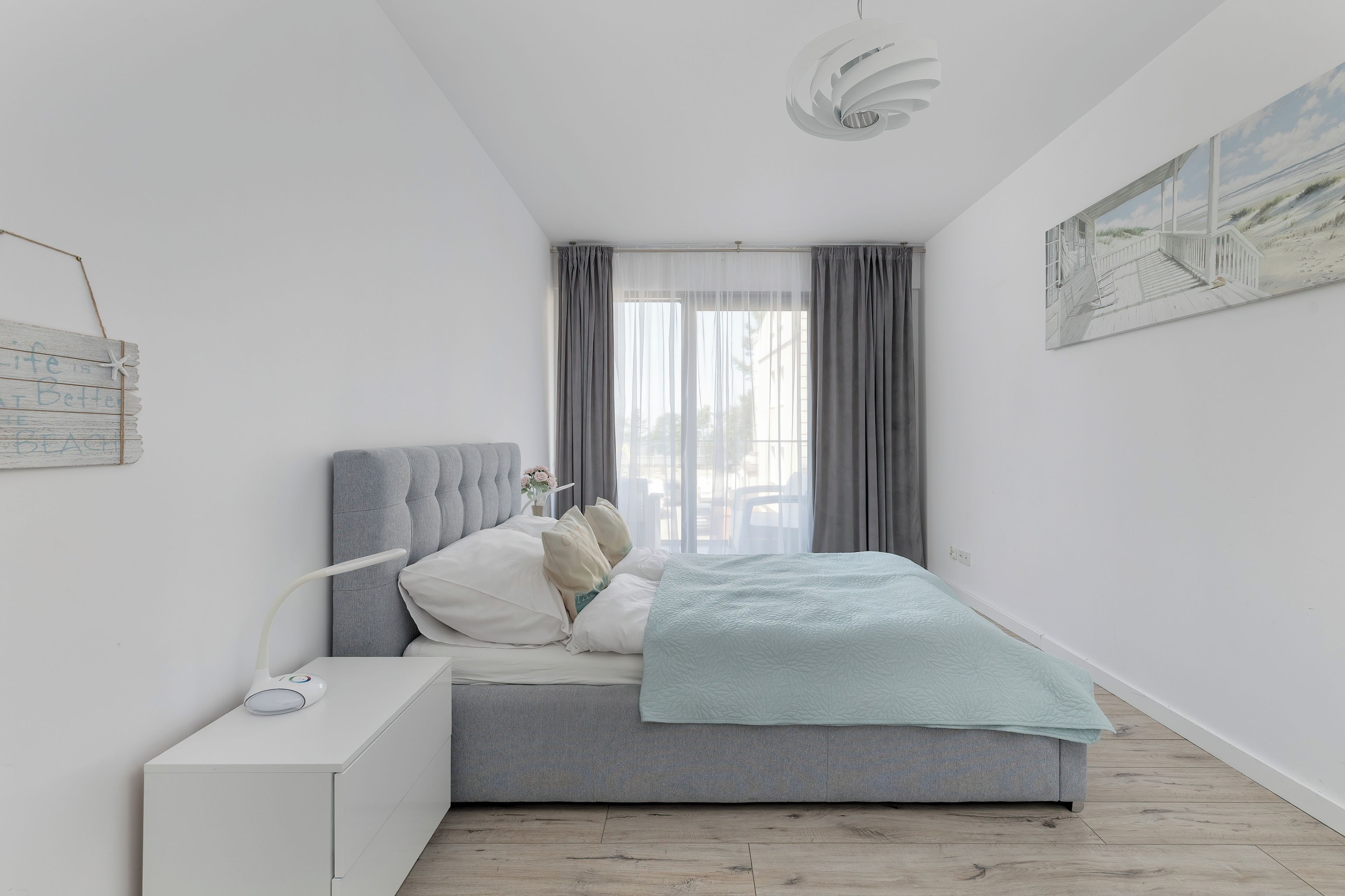 A bedroom with a bed, a bedside table, a lamp, a window with curtains, and a painting on the wall.
