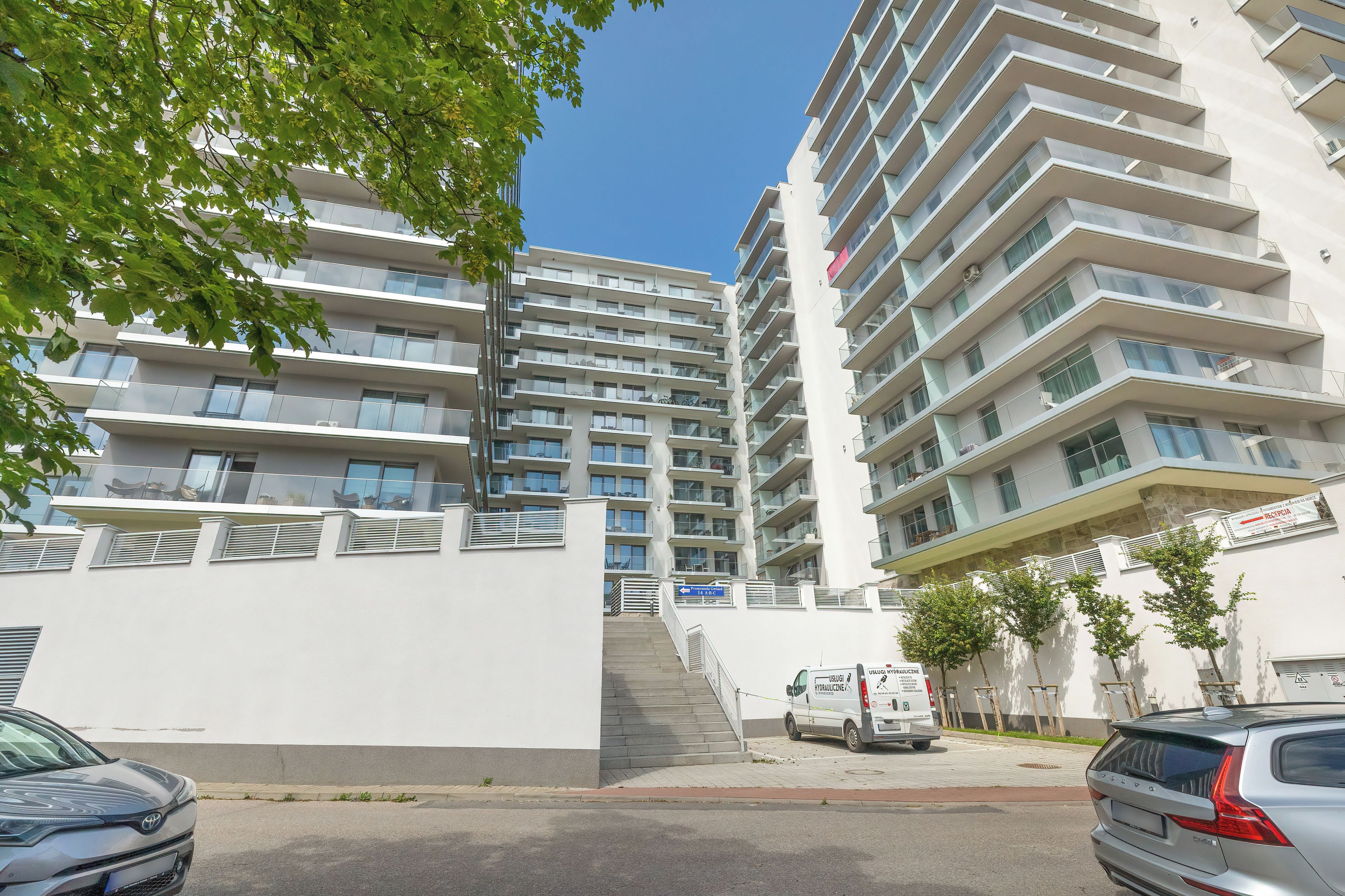 A multi-story residential building with balconies, a staircase leading to the entrance, and parked cars.
