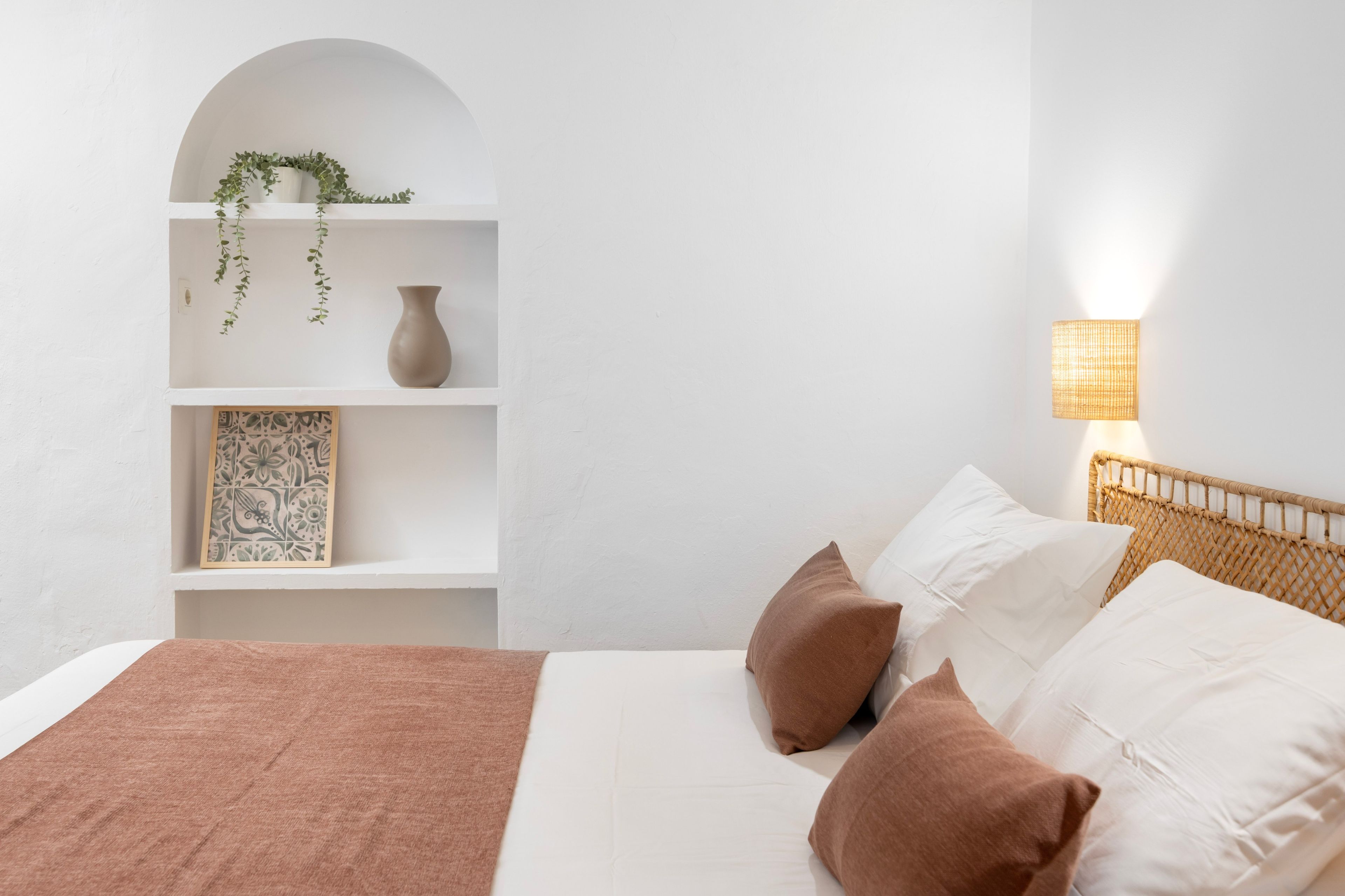 A bedroom with a bed, pillows, a bedside lamp, a shelf with a vase and a framed picture.