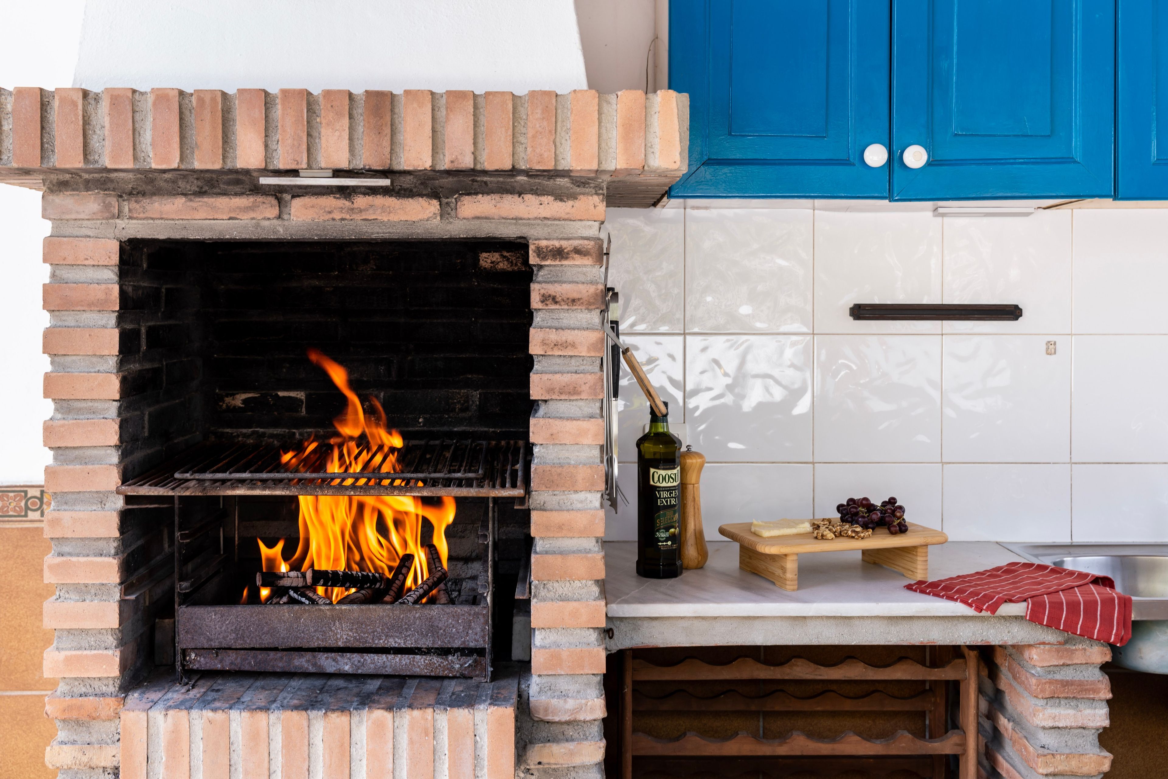 A brick-built outdoor grill with flames, a bottle of olive oil, a cutting board with nuts and grapes, and a red and white striped cloth.