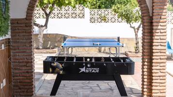 Une table de baby-foot de la marque « Premier Star », installée à l’extérieur, avec des murs en briques et une clôture blanche en treillis en arrière-plan.