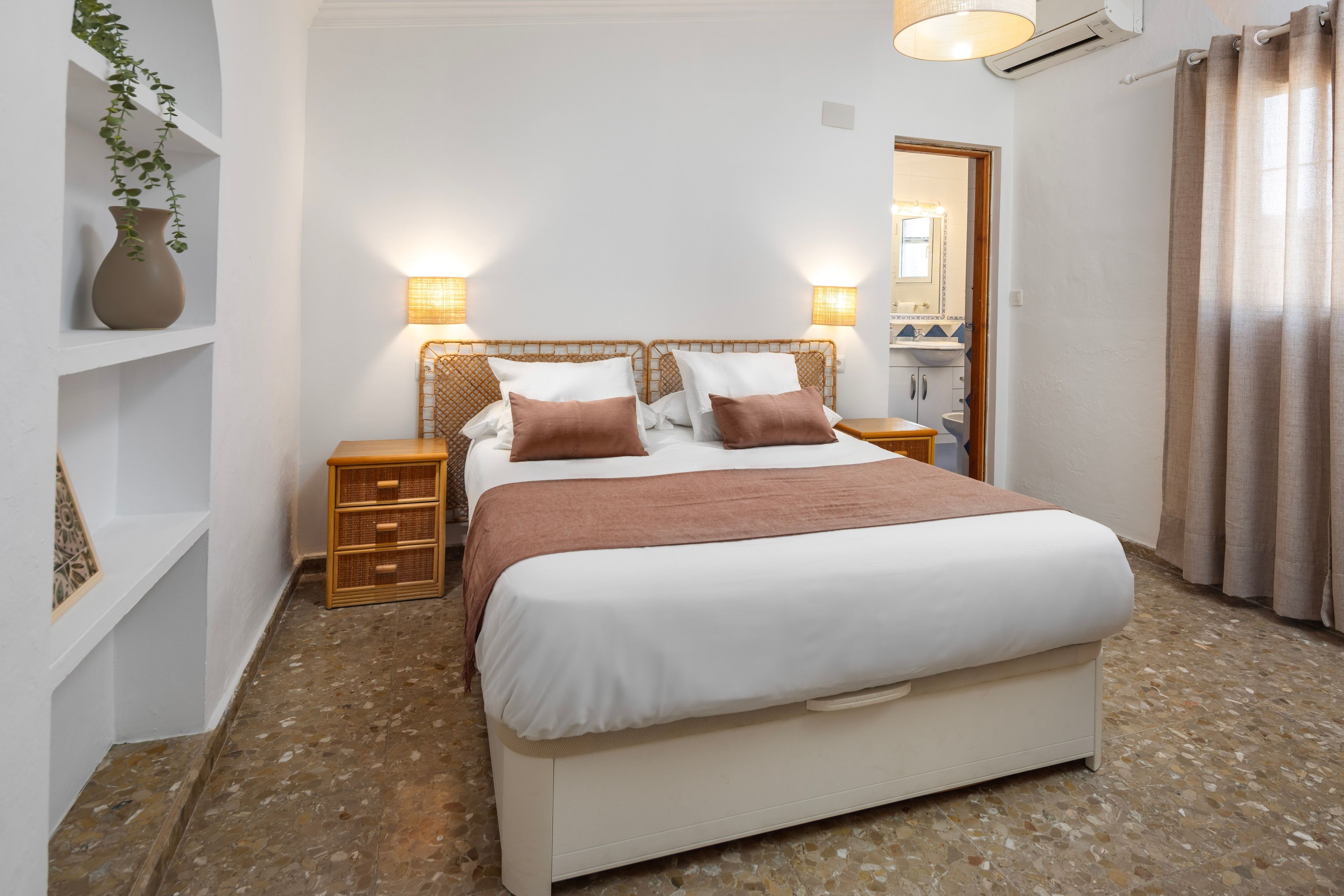 Ein Schlafzimmer mit Bett, Nachttischen, einer Wandlampe, einem Topfpflanzen und einem durchgegangenen Tür erkennbaren Badezimmer.