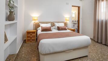 Ein Schlafzimmer mit Bett, Nachttischen, einer Wandlampe, einem Topfpflanzen und einem durchgegangenen Tür erkennbaren Badezimmer.
