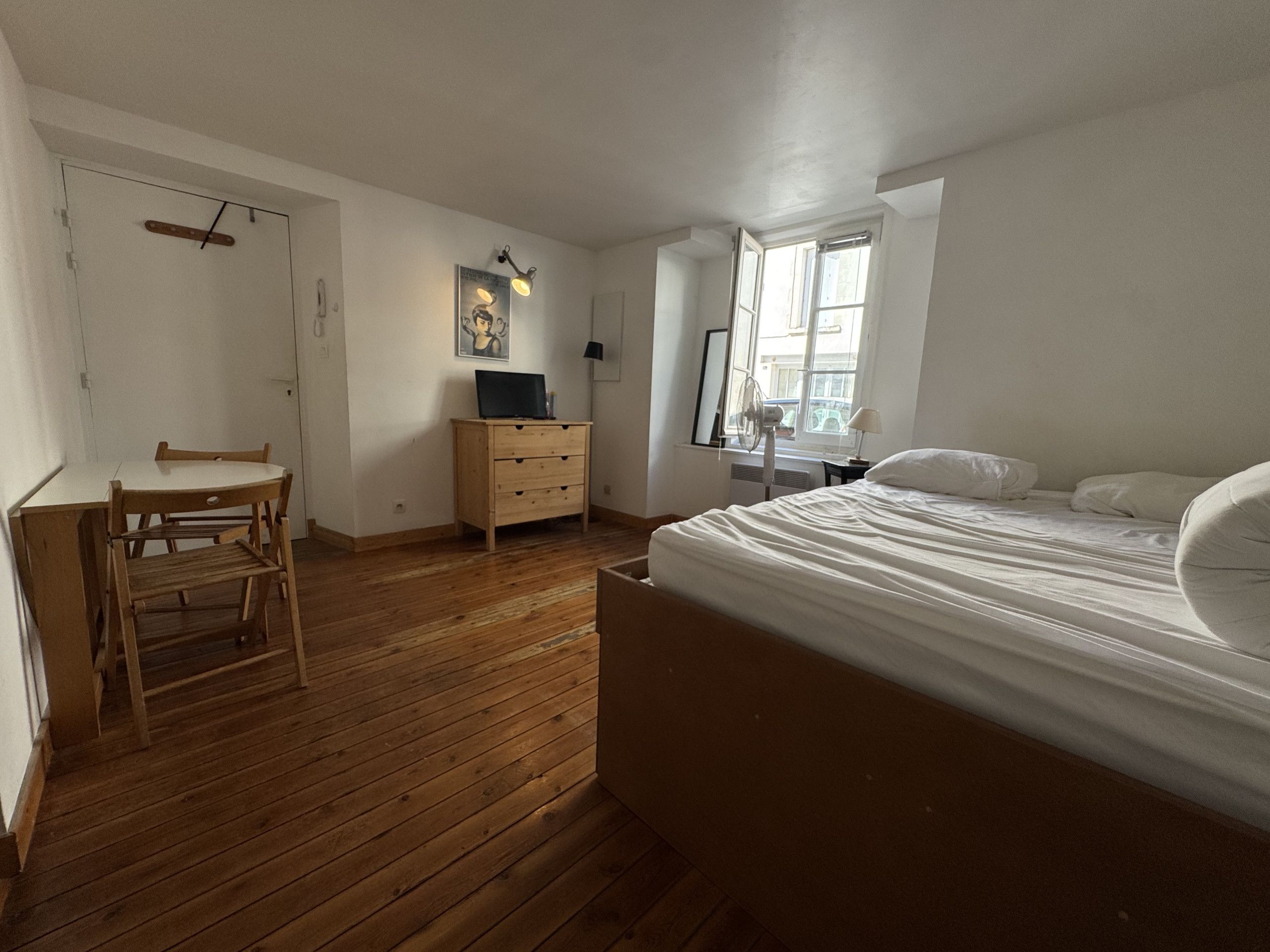 A bedroom with a wooden floor, a bed with white bedding, a desk with a chair, a dresser, and a window with a view.