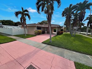 A residential property with a paved driveway, palm trees, and a house with a tiled roof. - 3BR Hallandale Beach House, Fenced Yard & BBQ, Sleeps 8, Free Parking (Hallandale Beach)