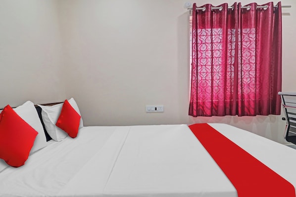 Cama com roupa de cama branca e detalhes vermelhos, uma tomada na parede e uma janela com cortinas vermelhas.