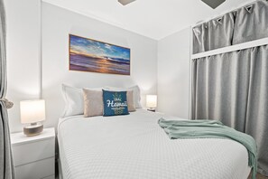 A hotel room with a bed, a nightstand, a lamp, a painting of a sunset, and a window with curtains. - Fully renovated condo 1 block from beach in Heart of Waikiki! Free parking! (Honolulu)