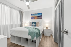 A bedroom with a bed, a nightstand, a wall-mounted lamp, a ceiling fan, and a painting of a sunset. - Fully renovated condo 1 block from beach in Heart of Waikiki! Free parking! (Honolulu)