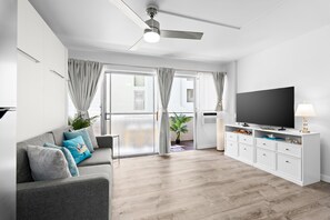 A modern living room with a grey sofa, a flat-screen TV, and a ceiling fan. - Fully renovated condo 1 block from beach in Heart of Waikiki! Free parking! (Honolulu)