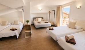Habitación de hotel con cuatro camas dispuestas en forma de U, una planta pequeña a la izquierda y un balcón con vistas.