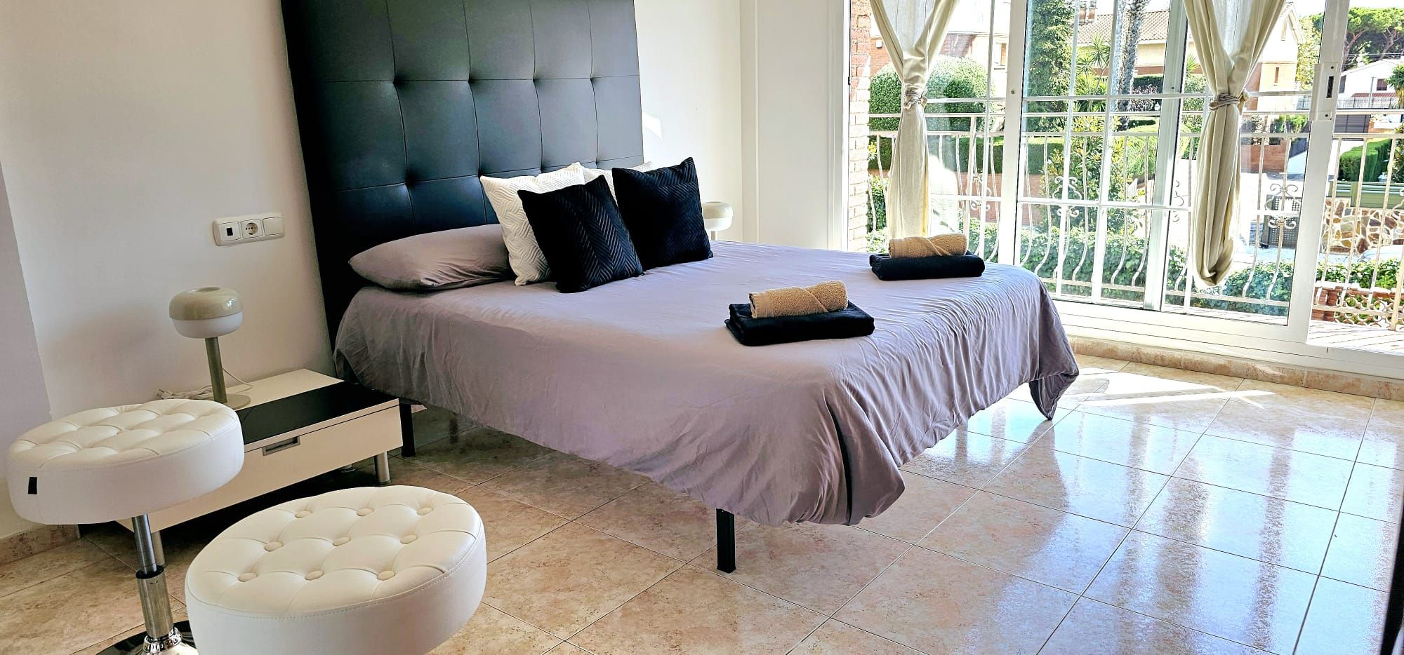 Ein modernes Schlafzimmer mit einem großen Bett, zwei weißen Hocker, einem Nachttisch und Blick auf einen Garten durch ein Fenster.