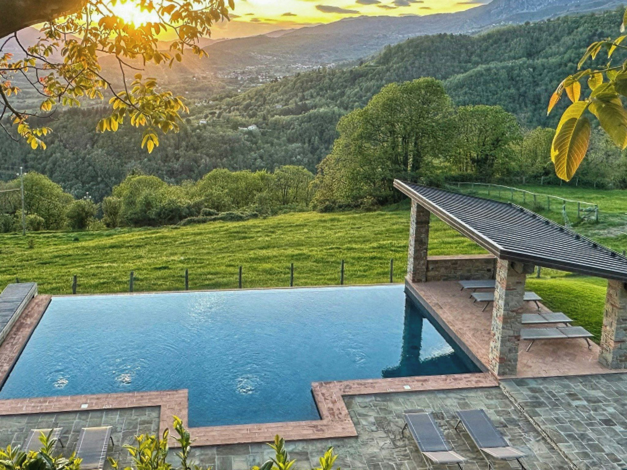Ein Schwimmbecken mit einer Steinterrasse, umgeben von einer Rasenfläche und Bäumen, mit Blick auf ein Tal und Berge im Hintergrund.
