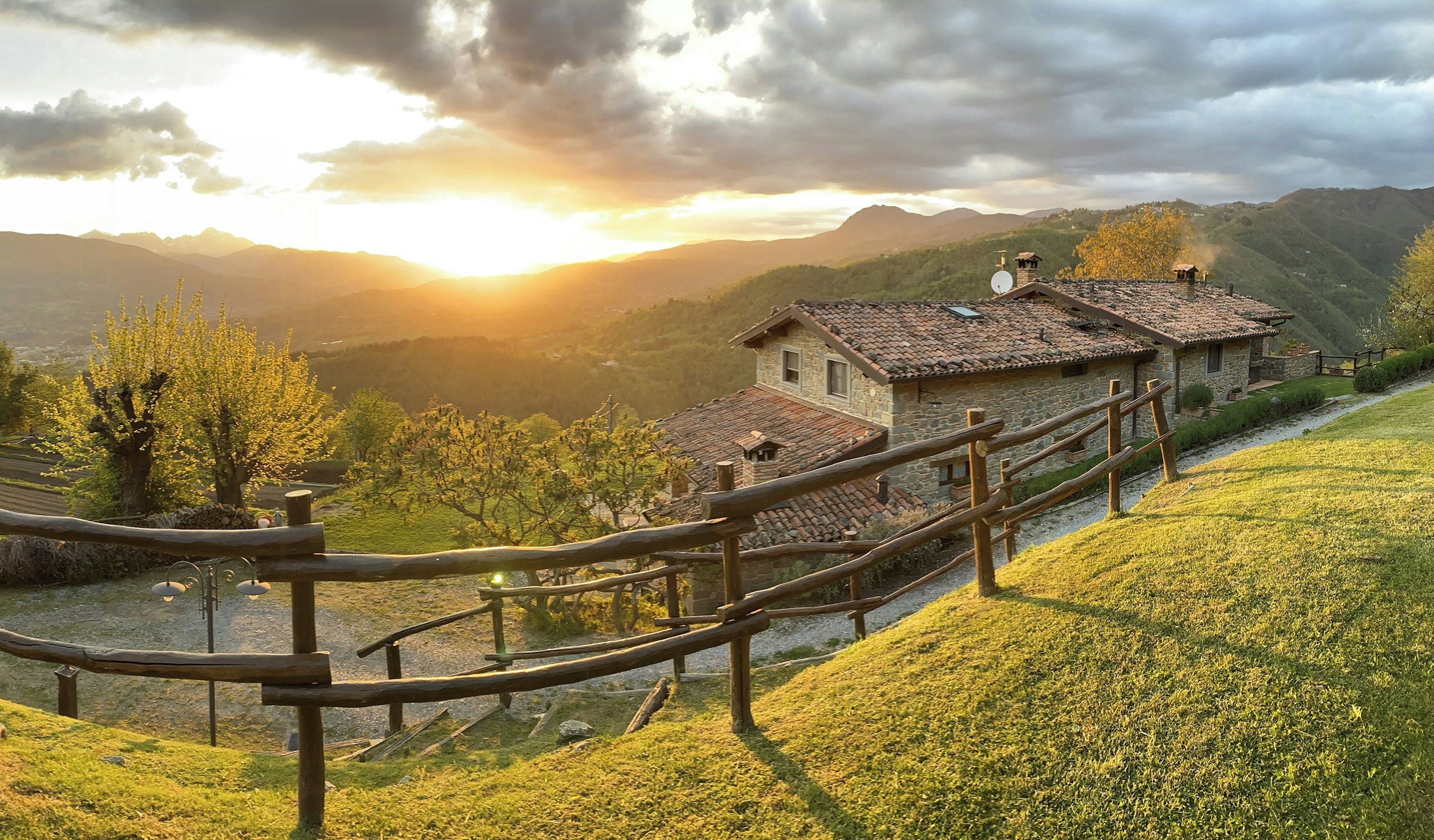 A stone house with a tiled roof, surrounded by a wooden fence, set against a backdrop of mountains and a sunrise.