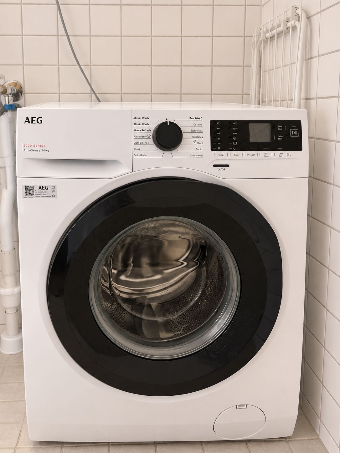 Een AEG 5000 serie wasmachine met voorlader en digita bedieningspaneel, in een witte betegelde badkamer.