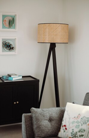 Eine Stehlampe mit gewebtem Schirm, eine schwarze Kommode mit zwei Türen und ein graues Sofa mit einem blumigen Kissen.