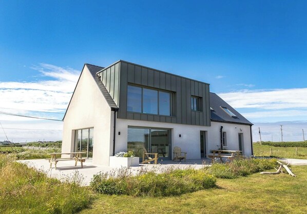 Une maison moderne à deux étages, dotée d’une grande baie vitrée, d’un patio avec des meubles d’extérieur et d’un toit en pente.