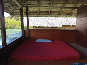 A single bed with a red patterned bedspread in a room with wooden walls and a thatched roof. - Cozzy Anno Homestay (Sawinggrai)
