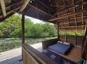 A wooden deck with a thatched roof structure, a bed with a blue and white striped bedspread, and a view of a lush green landscape.