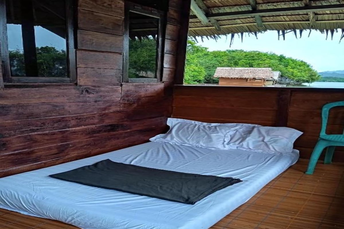 A single bed with white bedding and a dark blanket in a wooden room with a view of greenery.