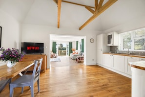 A spacious living area with a wooden floor, a dining table with chairs, a television displaying 'NETFLIX', and a view into the adjacent room through a doorway. - Chapel Cottage By My Getaways (Uckfield)