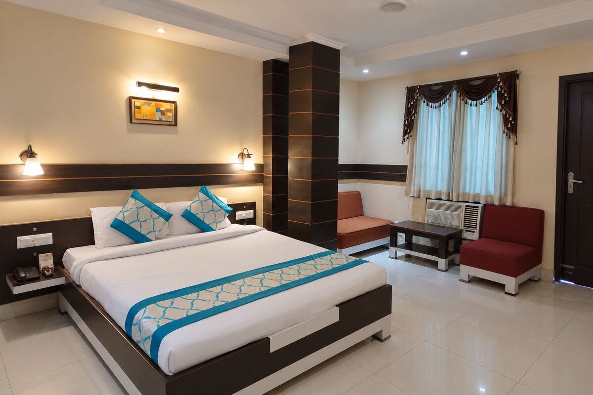 A hotel room with a large bed, bedside tables, a sofa, a chair, a small table, a heater, and a window with curtains.