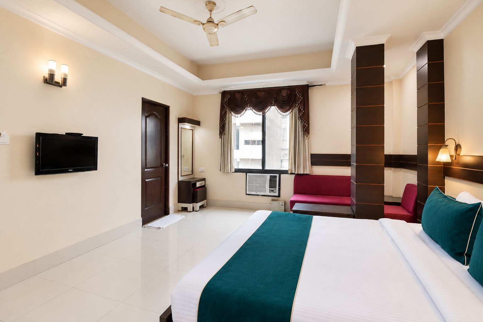 A hotel room with a bed, a television, a wardrobe, a mirror, a window with curtains, and a ceiling fan.