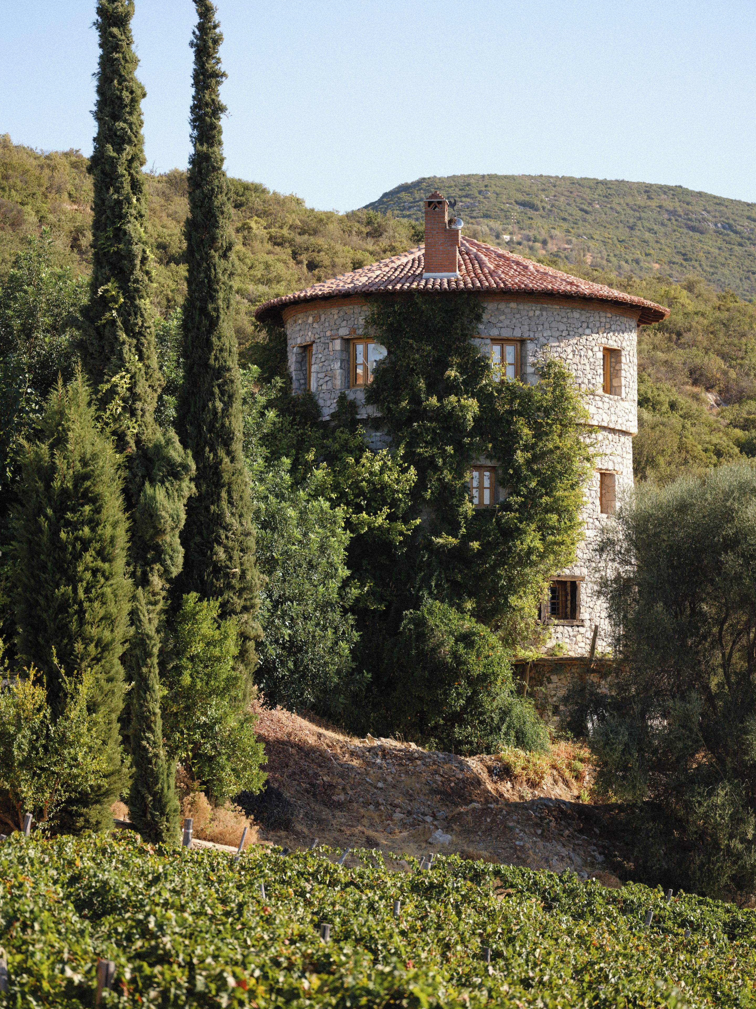 Un bâtiment en pierre, surmonté d’un toit de tuiles rouges, entouré de verdure et de collines.