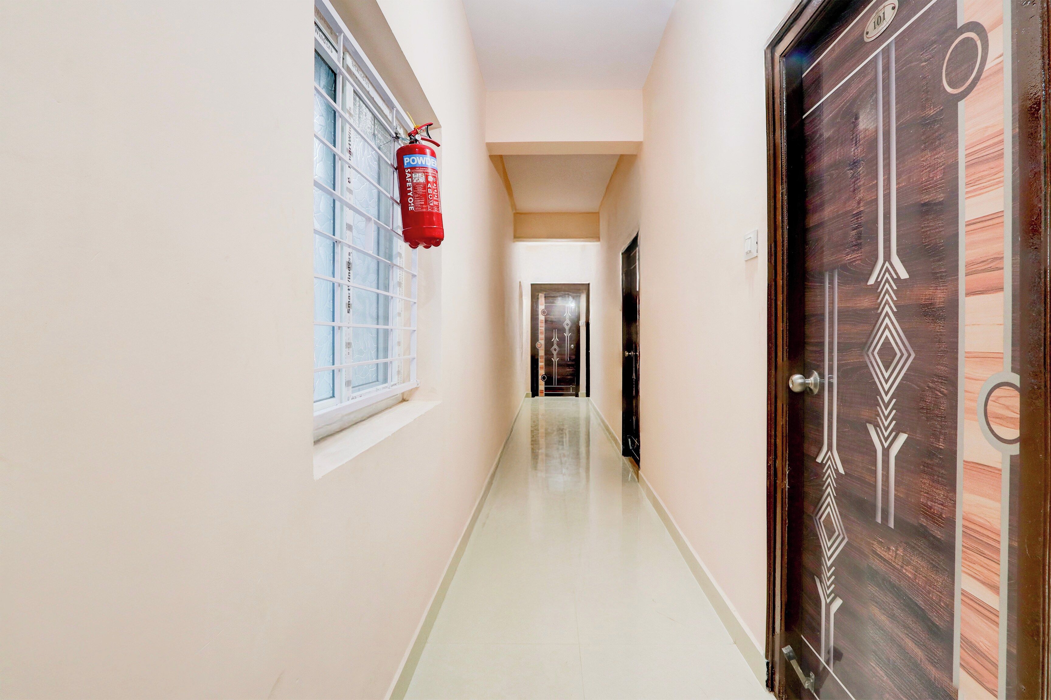 Un couloir avec un extincteur fixé au mur, une porte ornée d’un motif géométrique et une fenêtre à carreaux.