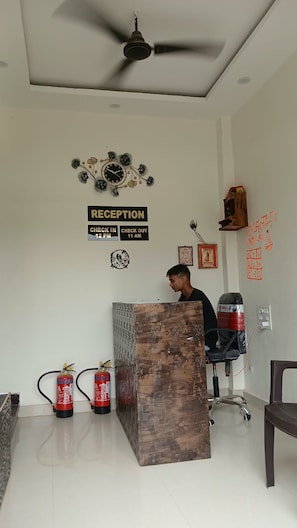 A reception area with a reception desk, ceiling fan, wall clock, fire extinguishers, and a wall-mounted sign indicating check-in and check-out times. - Kashi Prabha Homestay (Varanasi)