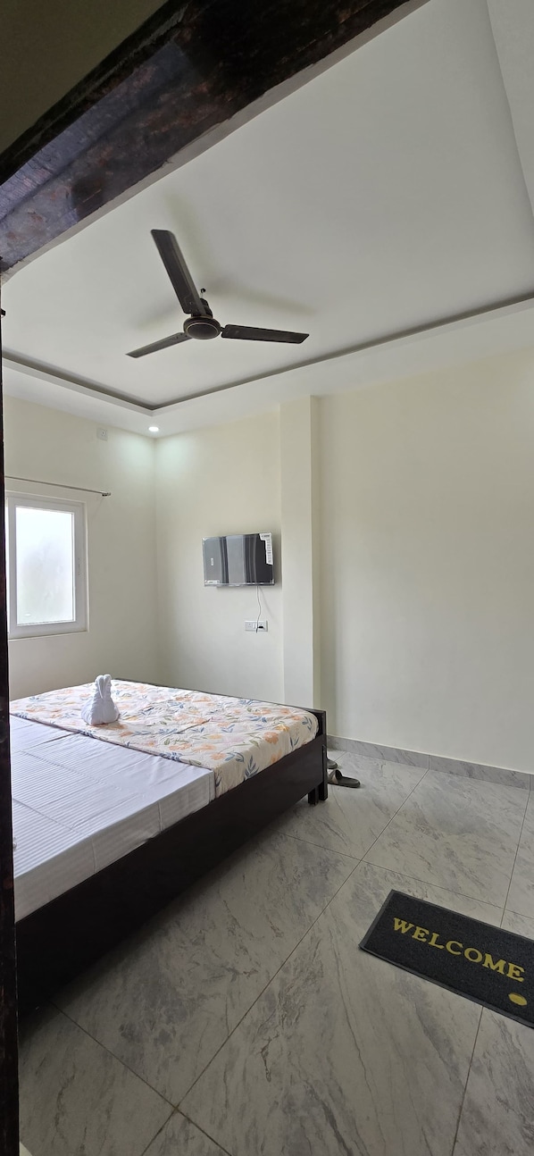 A bedroom with a bed, a ceiling fan, a TV mounted on the wall, and a welcome mat on the floor. - Kashi Prabha Homestay (Varanasi)