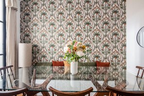 Een eethoek met een glazen tafel, houten stoelen, een vaas met bloemen en behang met een patroon.