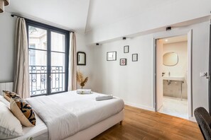A bedroom with a bed, a window with curtains, a door leading to a bathroom, and a wall with framed pictures. - Centrally Located 115SQM Residence In the Heart of Marais (Paris)