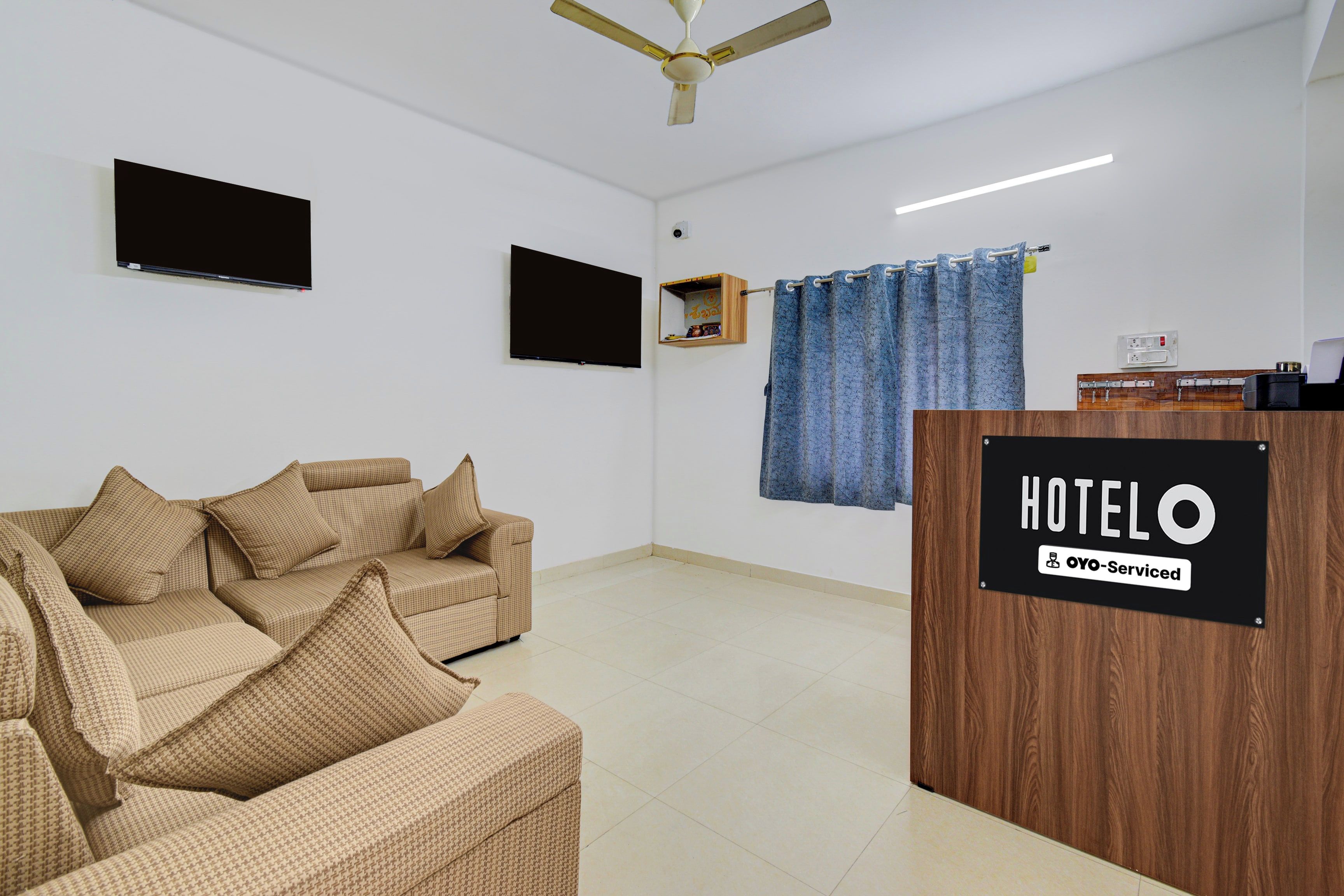 Habitación de hotel con sofá, televisor de pantalla plana, ventilador de techo, mostrador de recepción de madera y cortina azul.