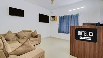 Habitación de hotel con sofá, televisor de pantalla plana, ventilador de techo, mostrador de recepción de madera y cortina azul.