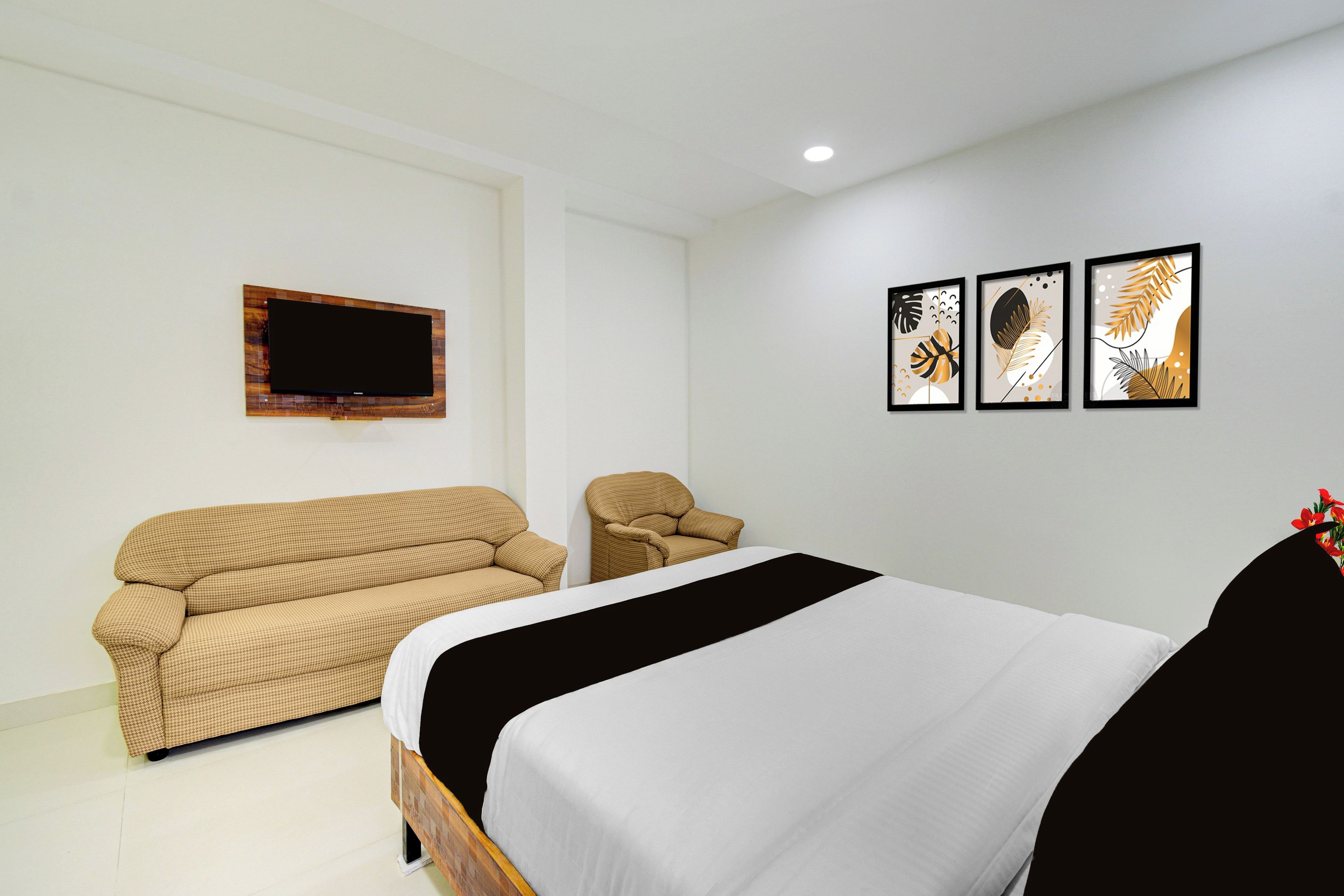 A hotel room with a bed, a sofa, a chair, a flat-screen TV mounted on the wall, and three framed artworks on the wall.