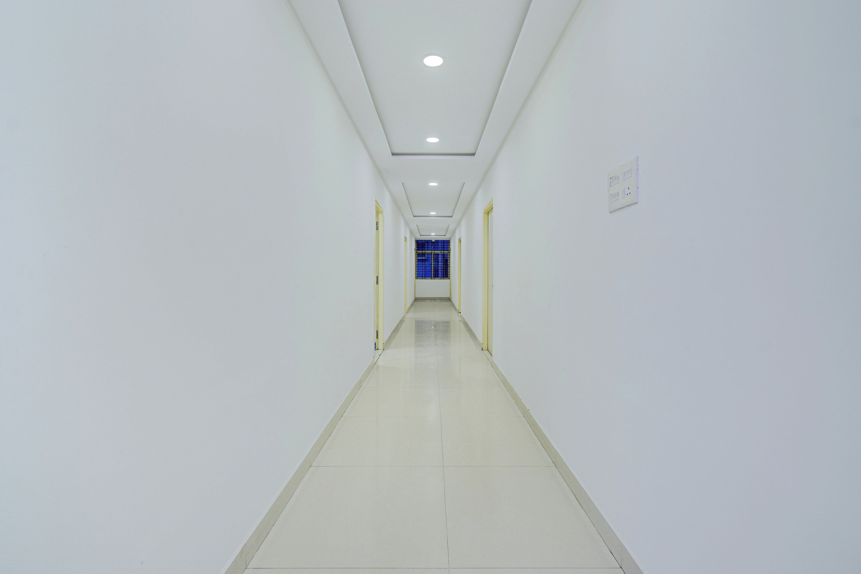 A long, narrow hallway with white walls and a tiled floor, leading to a room with a blue window.