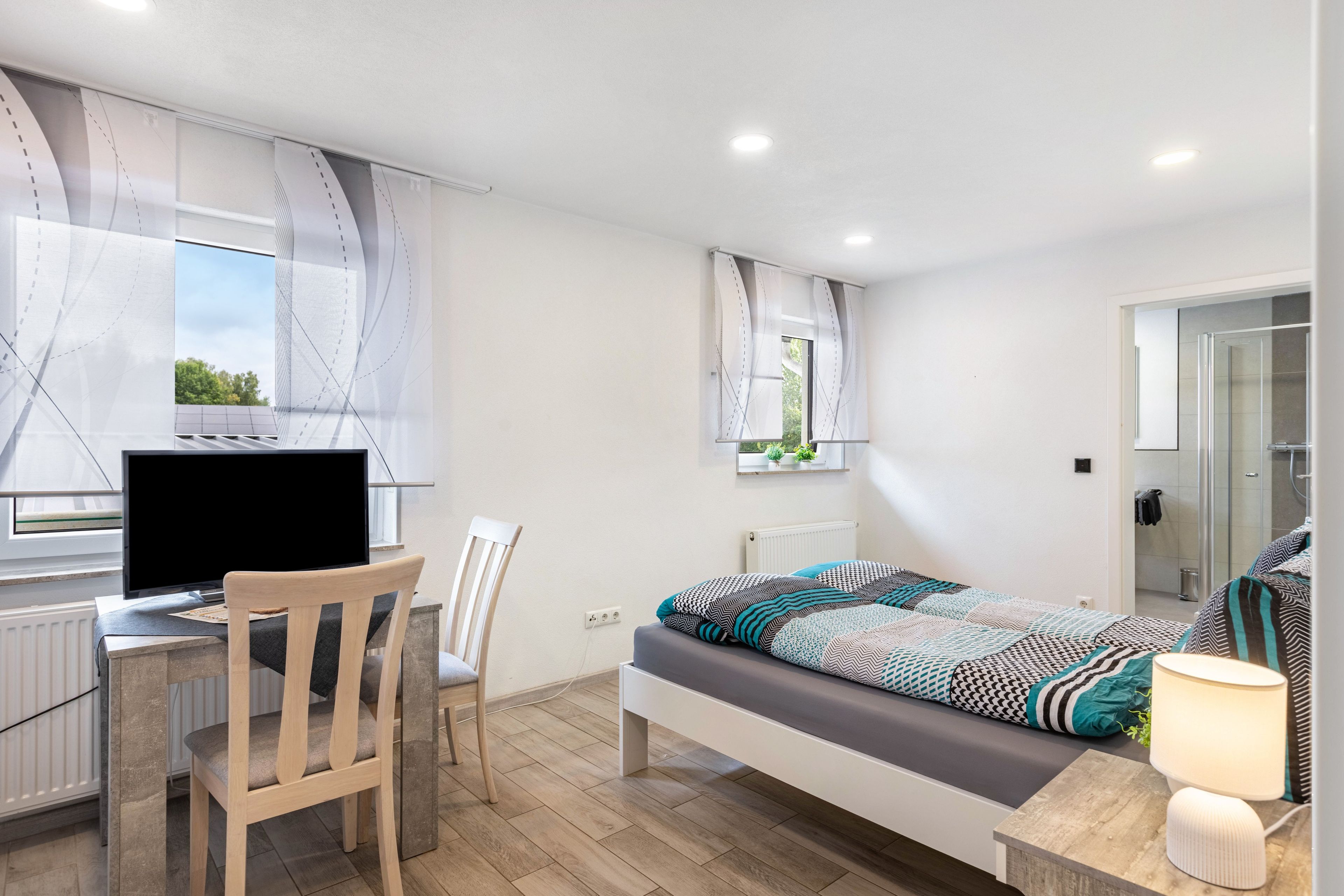 Ein Schlafzimmer mit einem Bett, einem Schreibtisch mit Computer, einem Stuhl und einem Fenster mit Vorhängen.