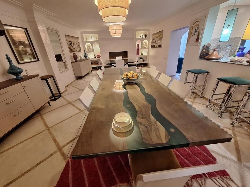 Un comedor con una mesa larga, sillas y un tapete rojo. Se observan taburetes de bar y una zona de cocina al fondo.