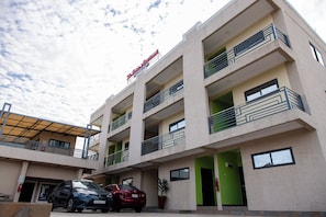 Un edificio de varios pisos con balcones, un estacionamiento con coches y un letrero que indica un negocio o servicio.