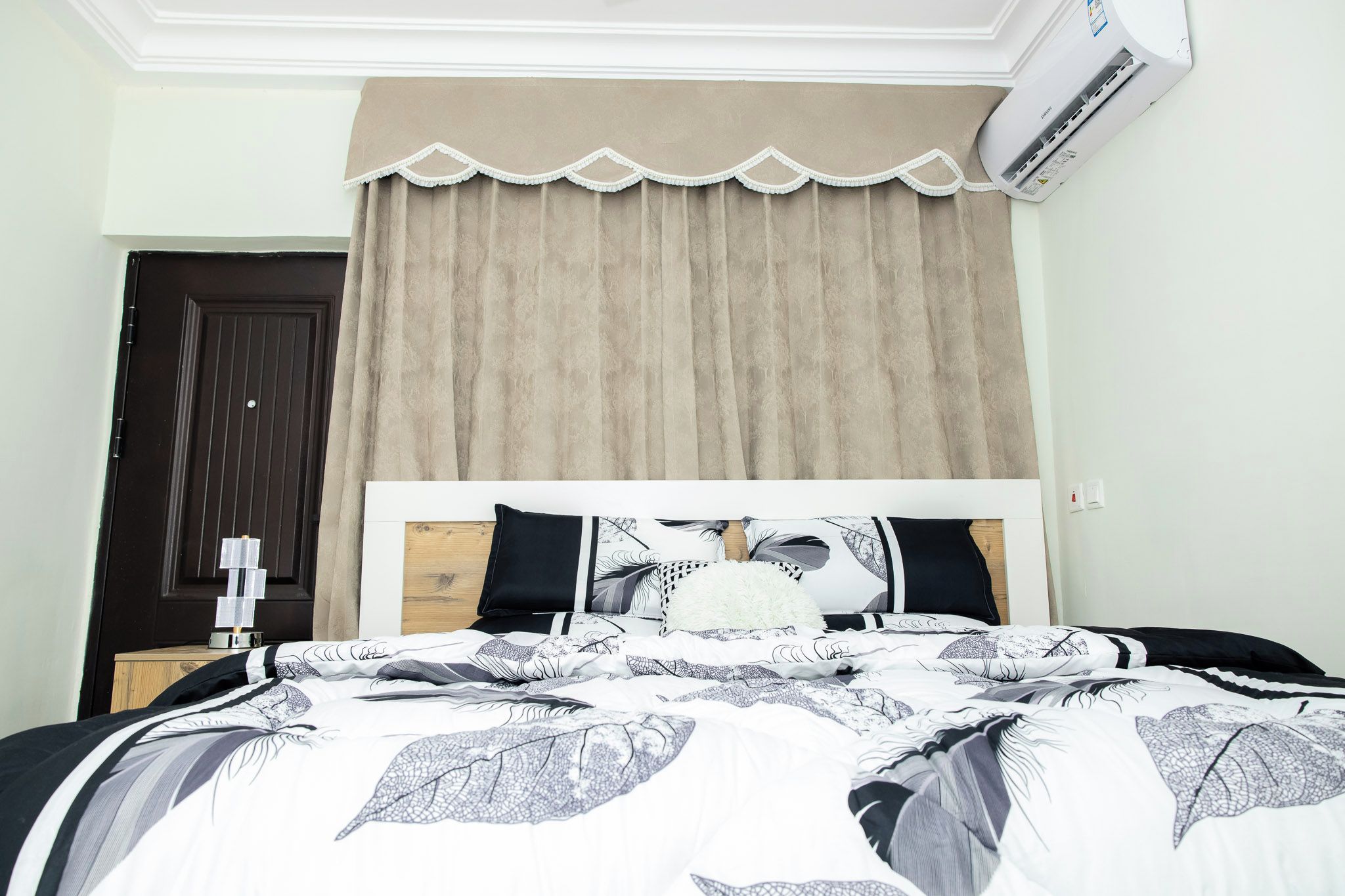 A bedroom with a bed, a window with beige curtains, a dark door, and an air conditioning unit.