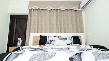 A bedroom with a bed, a window with beige curtains, a dark door, and an air conditioning unit.