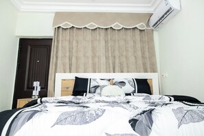 Een slaapkamer met een bed, een raam met beige gordijnen, een donkere deur en een airconditioner.