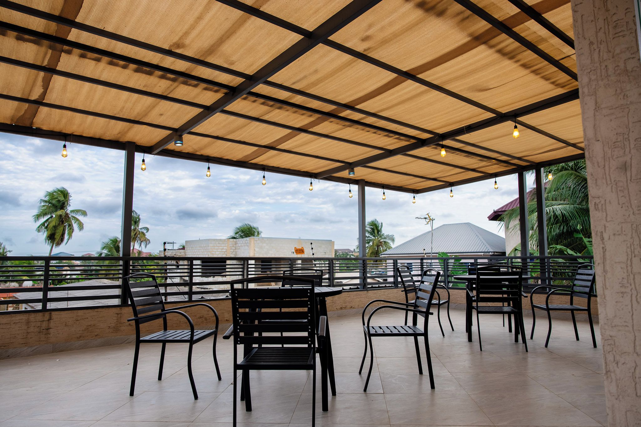 An outdoor seating area with tables and chairs, a covered roof, and a view of palm trees and buildings.