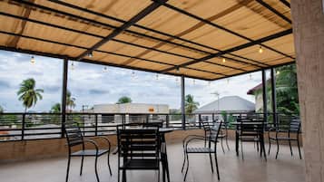 An outdoor seating area with tables and chairs, a covered roof, and a view of palm trees and buildings.