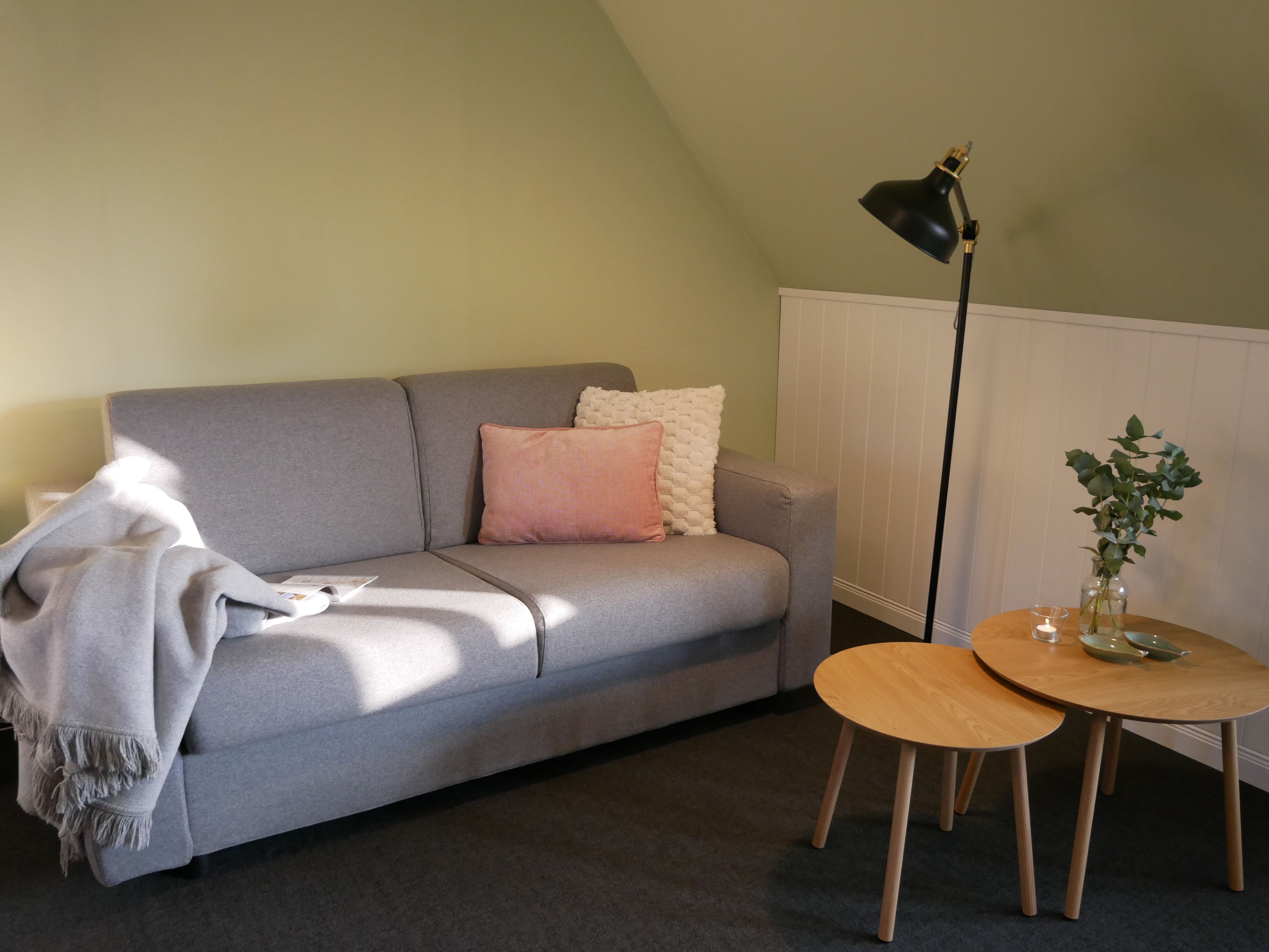 Ein graues Sofa mit einer Decke, ein kleiner Tisch mit einer Vase und eine Stehlampe in einem Zimmer mit schrägem Dach.