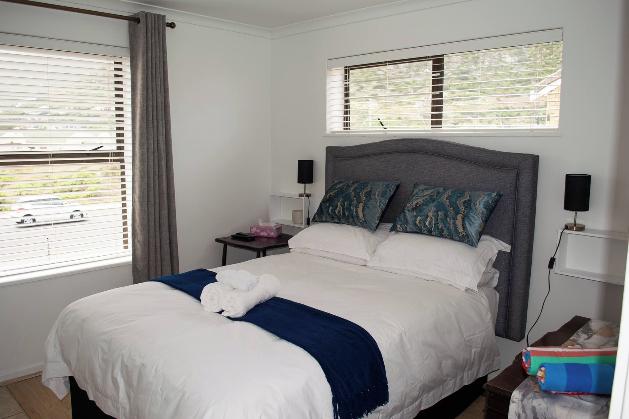 Quarto de hotel com uma cama grande, duas luminárias de cabeceira, mesa de apoio e uma janela com persianas.