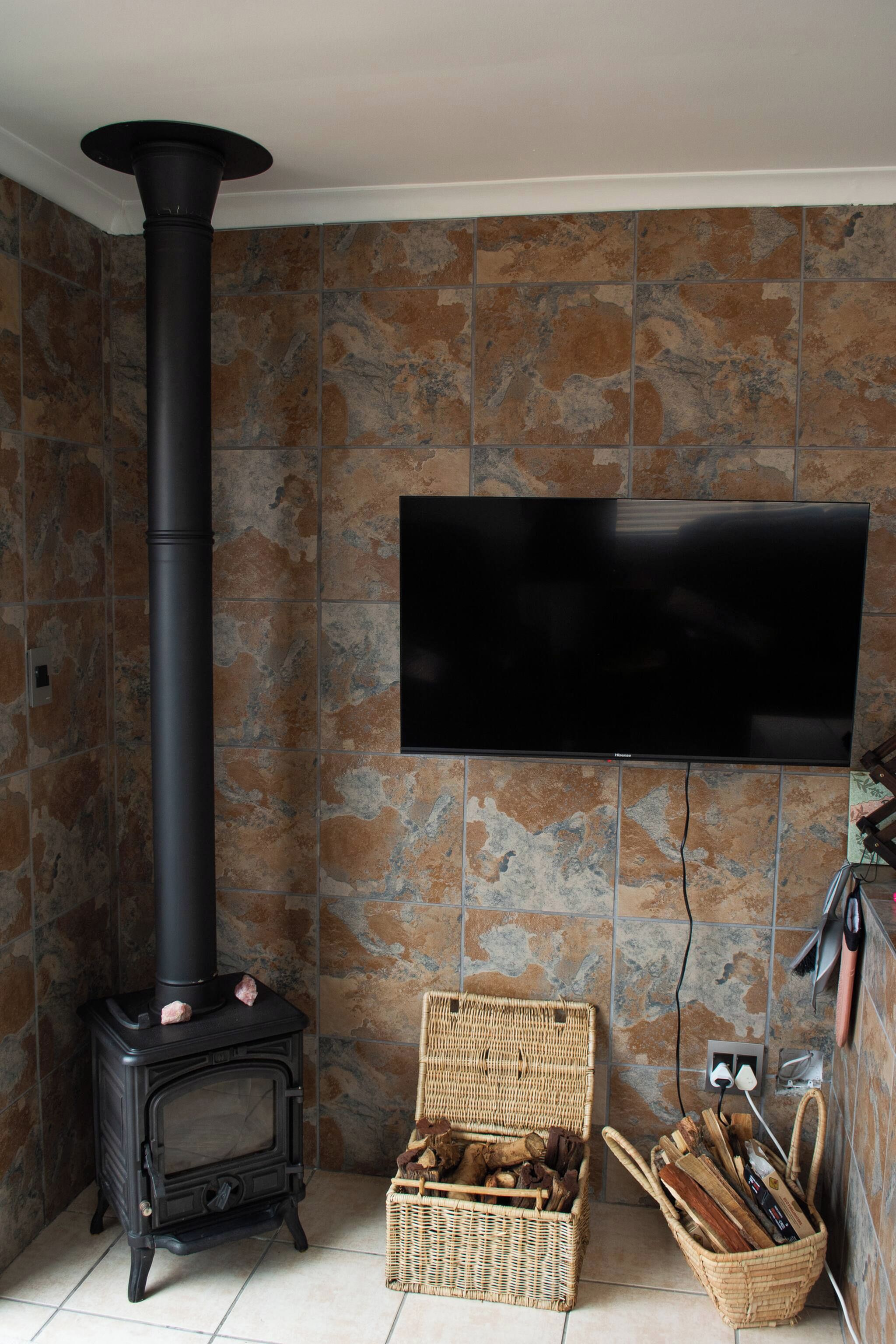 A room with a black wood-burning stove, a flat-screen TV mounted on the wall, and wicker baskets filled with firewood.